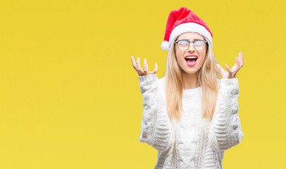 Young beautiful blonde woman wearing christmas hat over isolated background crazy and mad shouting and yelling with aggressive expression and arms raised. Frustration concept.