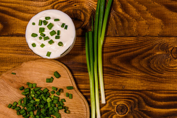 Glass bowl with sour cream and cutting board with chopped green onion on wooden table. Top view
