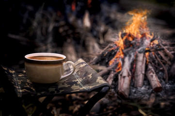 Cup of coffee on field chair with bonfire background.