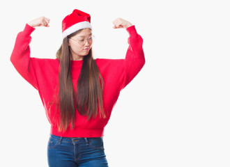 Young Chinese woman over isolated background wearing christmas hat showing arms muscles smiling proud. Fitness concept.