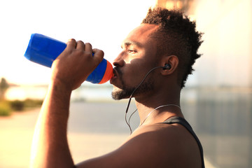 African man in sports clothing drinking water while standing outside, at sunset or sunrise. Runner.