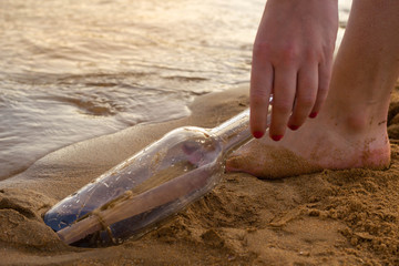 Message in a bottle on the beach, with a letter of distress in the sand on the beach in the summer and warm colors. The female hand takes out bottle with a letter from sea.