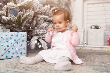 Small girl in the pink pajama sitting near the christmas tree playing with the garland