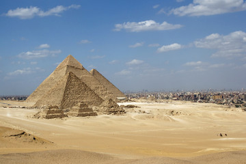 Fototapeta na wymiar Great Pyramids of Giza set against a bright blue sky and golden yellow desert sands but with the encroaching city of Cairo in the background