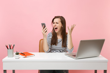 Young aggressive woman spreading hands screaming talking on mobile phone while sit working at office with pc laptop isolated on pastel pink background. Achievement business career concept. Copy space.