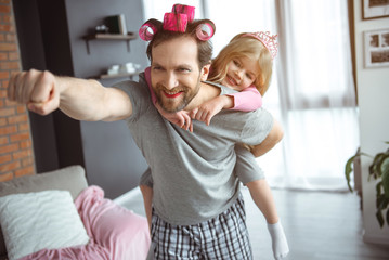 I am superman. Portrait of happy father is carrying his daughter on back and laughing. He is stretching hand forward. Man has make-up on face and hair curlers  - obrazy, fototapety, plakaty