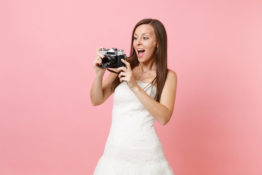 Amazed bride woman in wedding dress taking pictures on retro vintage photo camera choosing staff, photographer isolated on pink background. Wedding to do list. Organization of celebration. Copy space.