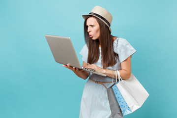 Portrait of young woman in summer dress, straw hat holding packages bags with purchases after online shopping, using laptop pc computer isolated on blue pastel background. Copy space for advertisement