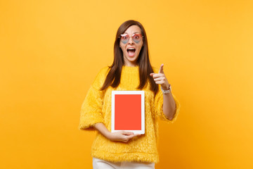 Amazed woman in heart glasses pointing index finger on camera, holding tablet pc computer with blank black empty screen isolated on bright yellow background. People sincere emotions lifestyle concept.
