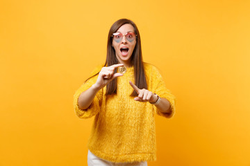 Excited young woman in heart glasses pointing index finger on bitcoin, metal coin of golden color, future currency isolated on yellow background. People sincere emotions, lifestyle. Advertising area.