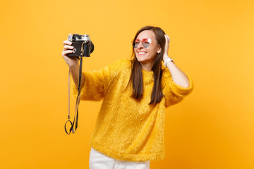 Funny young woman in heart eyeglasses doing taking selfie shot on retro vintage photo camera, putting hand on head isolated on yellow background. People sincere emotions, lifestyle. Advertising area.