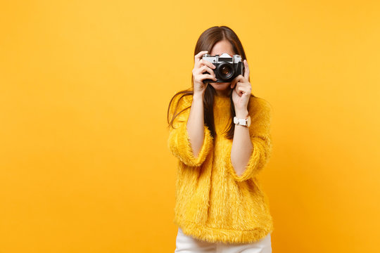 Brunette young woman in fur sweater covering face and taking pictures on retro vintage photo camera isolated on bright yellow background. People sincere emotions, lifestyle concept. Advertising area.