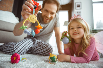 Seeing by the eyes of infant. Low angle of happy dad and small sister holding playthings in front of the toddler. They are relaxing on soft carpet and laughing  