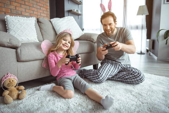 Family entertainment. Full length portrait of happy girl using joystick while looking forward with excitement. Her dad is sitting on floor and smiling 