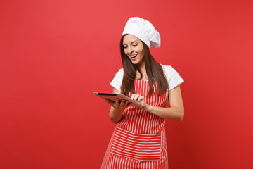 Housewife female chef cook or baker in striped apron, white t-shirt, toque chefs hat isolated on red wall background. Fun housekeeper woman looking for recipe in tablet pc. Mock up copy space concept.