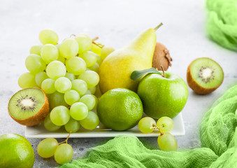 Fresh raw organic green toned fruit on white ceramic square plate on stone kitchen table background. Pear and grapes with kiwi and lime and apples.