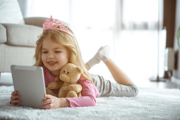 Portrait of happy little girl is watching interesting cartoon with excitement. She is holding tablet while lying on soft rug. Cute princess is smiling  © Yakobchuk Olena
