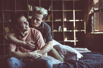 Toned portrait of handsome guy with dyed hair hugging boyfriend from behind. Wall unit with books on background