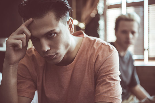 Close up portrait of dark-haired handsome guy looking away with unhappy expression and touching forehead. His boyfriend sitting behind on blurred background