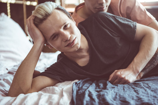 Happy relationship. Toned portrait of smiling young man with dyed hair lying in bed while boyfriend hugging him from behind