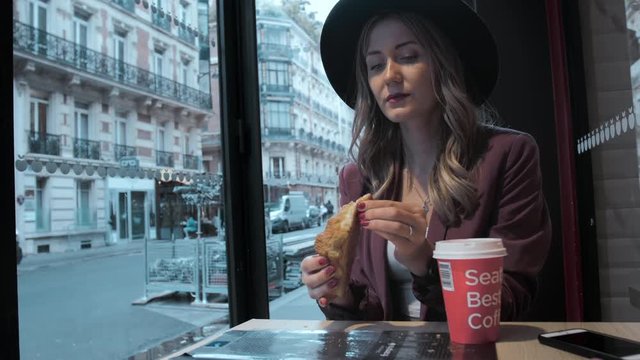 Beautiful French woman in a hat eats a croissant in a coffee shop on the background of a French street. On the table is a paper cup of coffee. Slow Motion.