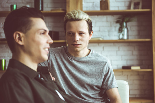 Toned portrait of handsome guy sitting on couch and gazing at dark-haired smiling friend. Focus on blond gentleman
