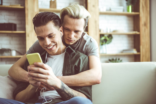 Toned waist up portrait of dark-haired guy sitting on couch and holding cellphone while his boyfriend hugging him from behind