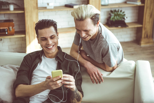 Toned waist up portrait of dark-haired young man sitting on couch and holding cellphone while his boyfriend standing behind