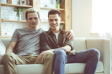 We are happy. Toned portrait of gay couple sitting on white couch and cuddling. Guys looking at camera with happy smiles