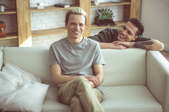 Look at me. Toned portrait of blond handsome guy sitting on couch while his boyfriend standing behind and gazing at him with smile
