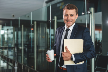 Waist up portrait of happy businessman holding paper cup and folder. He looking at camera with bright smile. Copy space on left side