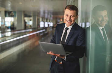 Waist up portrait of joyous man using laptop while leaning against wall. He looking at camera with...