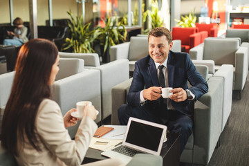 Adult businessman sitting afore female opponent in cafe and having hot drink. They looking at each other with smile