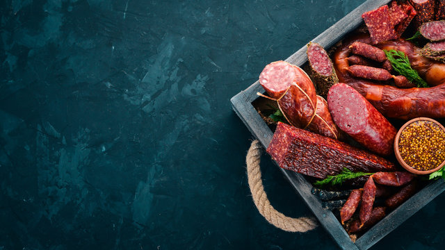 Assortment of sausages and snacks in a wooden box. Sausage Fuet, salami, paperoni. On a black background. Top view. Free space for your text.