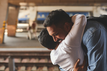 Positive smiling hindu man standing at the railway platform while embracing his little son