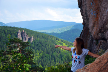 Naklejka premium A girl stands on a high rock on the edge of the cliff. Beautiful autumn forest. Autumn in the mountains. The girl looks dreamily into the distance. Unity with nature. Beautiful view from the top.