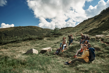 Happy five tourists with rucksacks walking to mount top together. Four women and man sitting on...