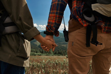 Close up of male and female while hand-holding with tenderness. They standing and carrying rucksacks while trekking among green mountains