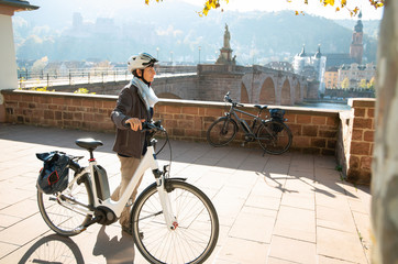 Frau mit E-Bike,  in herbstlichem Gegenlicht, vor Heidelberger  Altstadt mit Schloß und alter Brücke - obrazy, fototapety, plakaty
