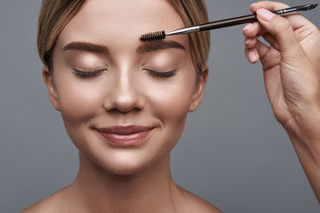Fototapeta premium Calm relaxed young lady sitting with her eyes closed and smiling while professional beautician brushing her eyebrows
