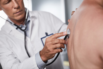 Breathe, do not breathe. Adult doctor in white robe making listening to lungs of man while using...