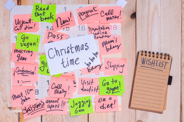 Christmas time reminder, written on a white paper note stuck to a busy full of green and pink sticky notes wall calendar on wooden background, notepad with Wish List on it