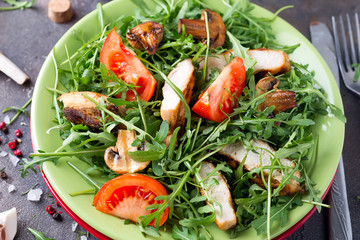 Fresh salad with chicken breast, arugula, mushrooms and tomatoes on green ceramic plate in a stone table.