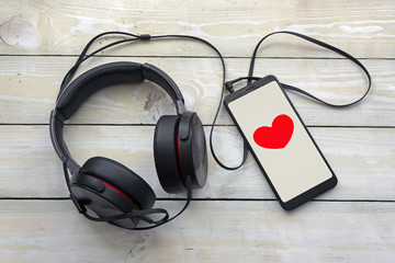 black headphones with smartphone on white wooden background, on the smartphone screen love symbol in the form of a heart top view