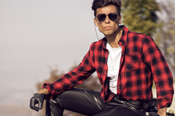 A young guy in a red shirt and leather pants sits and looks into the distance