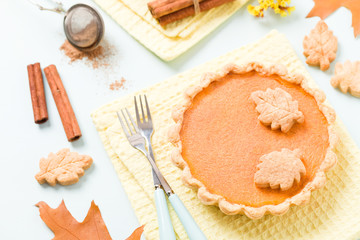 Pumpkin pie with cinnamon and cookies on yellow napkins on pastel blue background with autumn yellow leaves.