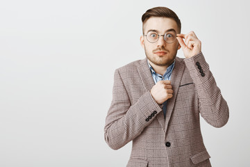 Waist-up shot of cute shy and insecure male nerd in glasses and fancy jacket holding fist near body popping eyes touching rim of eyewear standing unconfident and worried over gray wall