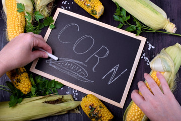 Raw and grilled maize cobs are framing the blackboard with the word "Corn" written with a piece of chalk