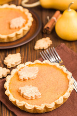 Pumpkin pie with cinnamon and cookies on brown napkins on wooden background with autumn yellow leaves.