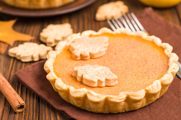 Pumpkin pie with cinnamon and cookies on brown napkins on wooden background with autumn yellow leaves.